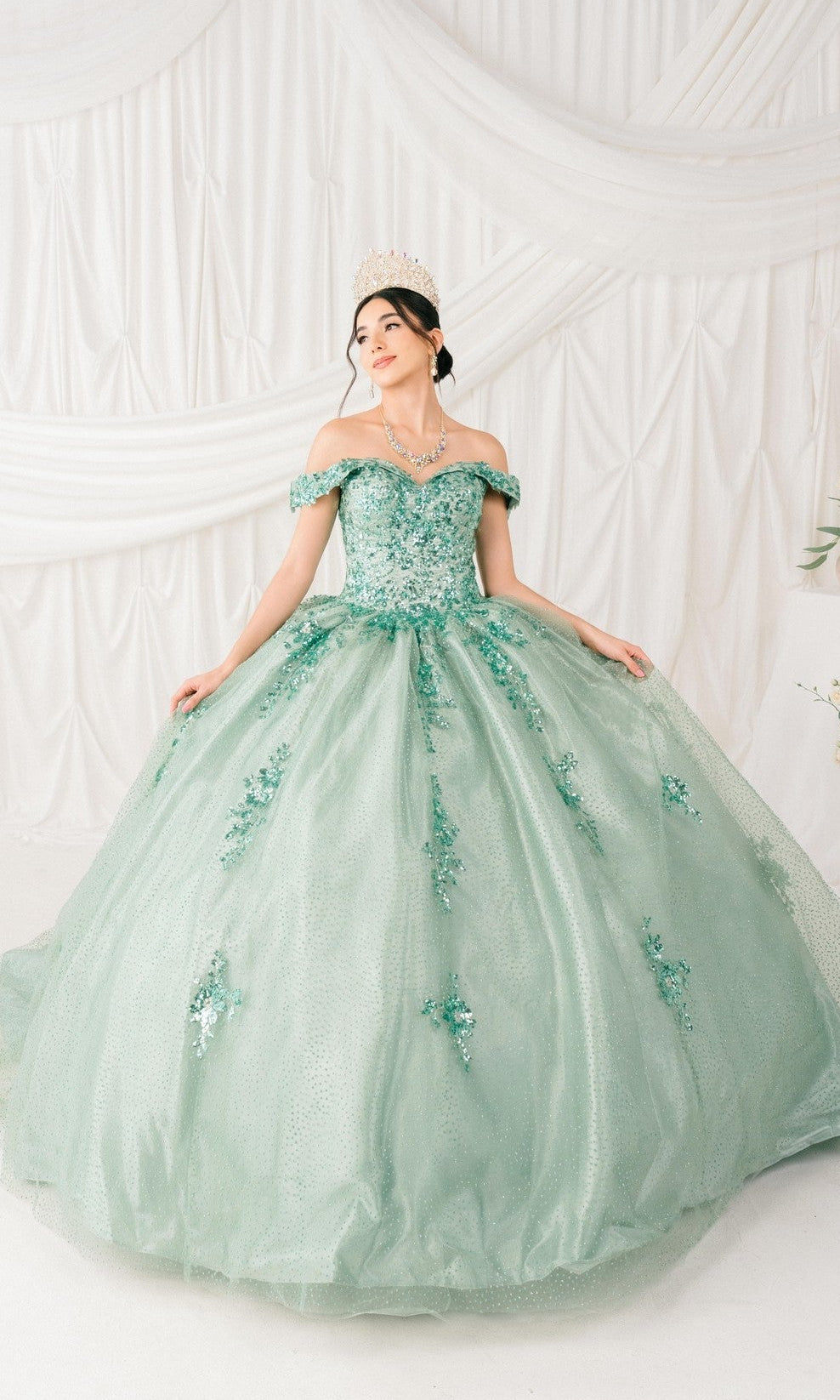 A woman in the Quinceanera Ball Gown 8060J, featuring an off-the-shoulder design, floral embellishments, and a lace-up back with a tiara, stands against a white draped backdrop.